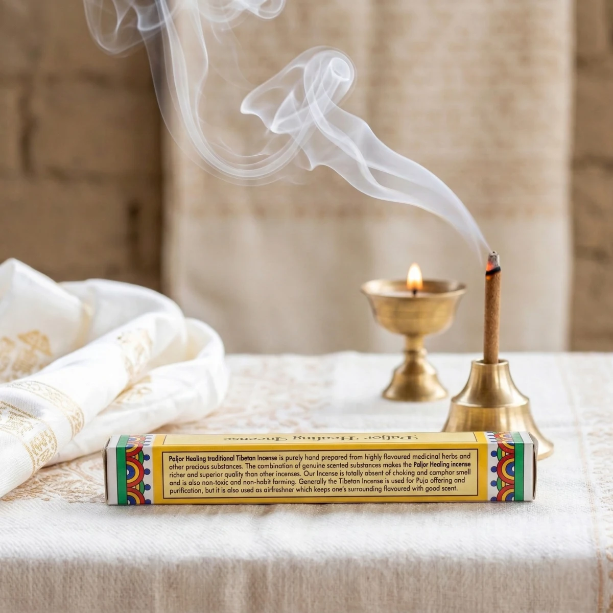 Paljor Healing Incense — Tibetan medicinal herbal stick burning in brass holder beside butter lamp, product box with mandala motif border